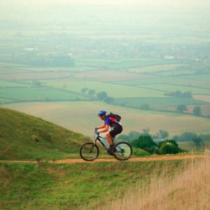 An image of an adult cycling on top of a hill, with a beautiful coutryside view behind them.