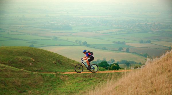 An image of an adult cycling on top of a hill, with a beautiful coutryside view behind them.